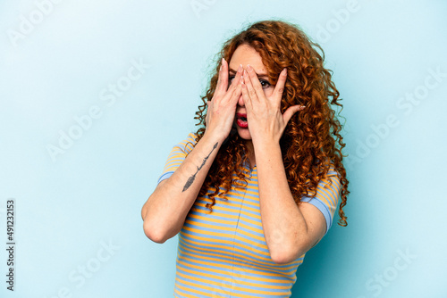 Young ginger caucasian woman isolated on blue background blink through fingers frightened and nervous.