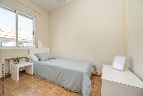 Single bed with blue bedspread with white headboard, matching side table and aluminum window with radiator below in bedroom with parquet floor © Toyakisfoto.photos