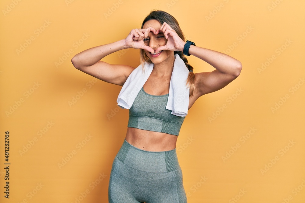 Beautiful hispanic woman wearing sportswear and towel doing heart shape with hand and fingers smiling looking through sign Beautiful hispanic woman wearing sportswear and towel doing heart shape with hand and fingers smiling looking through sign
