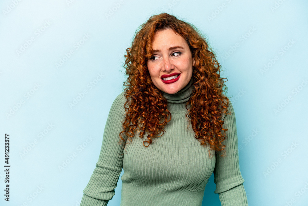 Young ginger caucasian woman isolated on blue background laughs and closes eyes, feels relaxed and happy. Young ginger caucasian woman isolated on blue background laughs and closes eyes, feels relaxed and happy.