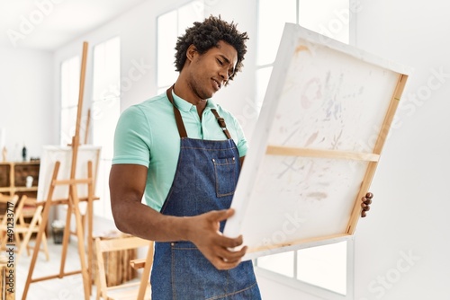 Young african american artist man with serious expression holding canvas at art studio.