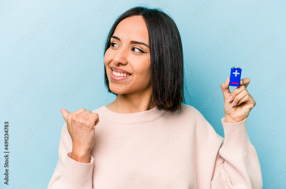 Young hispanic woman holding batteries isolated on blue background points with thumb finger away, laughing and carefree. Young hispanic woman holding batteries isolated on blue background points with thumb finger away, laughing and carefree.