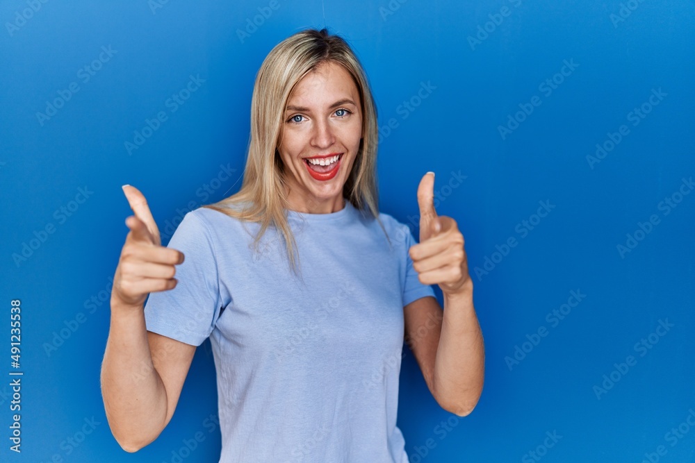 Beautiful blonde woman wearing casual t shirt over blue background pointing fingers to camera with happy and funny face. good energy and vibes. Beautiful blonde woman wearing casual t shirt over blue background pointing fingers to camera with happy and funny face. good energy and vibes.