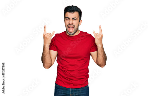 Hispanic man with beard wearing casual red t shirt shouting with crazy expression doing rock symbol with hands up. music star. heavy concept.