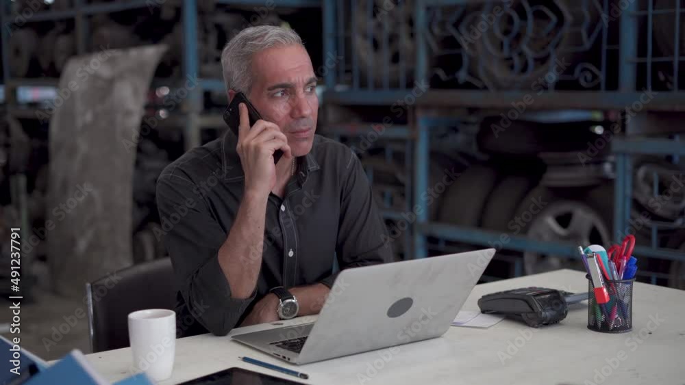 confident senior entrepreneur sitting on desk talking with mobile phone using laptop computer in warehouse of old used car engine . happy old businessman owner start up