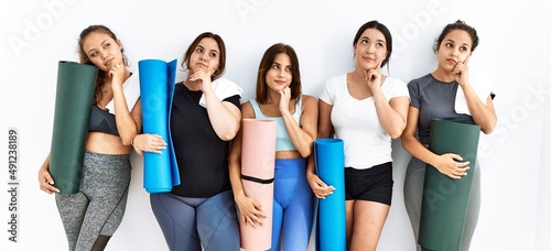 Group of women holding yoga mat standing over isolated background with hand on chin thinking about question  pensive expression. smiling with thoughtful face. doubt concept.