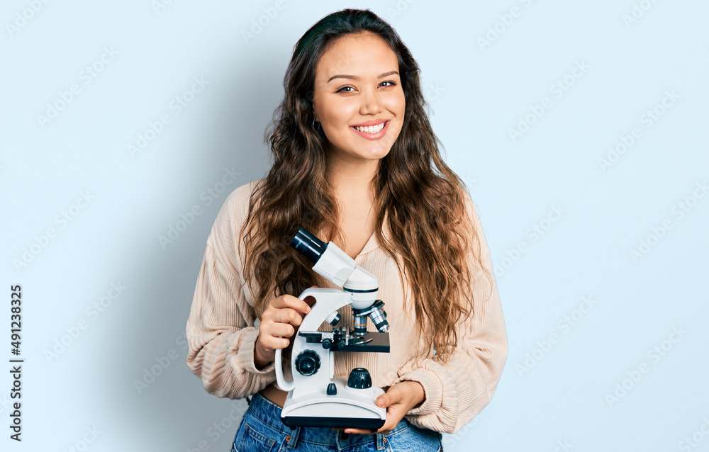 Young hispanic girl holding microscope looking positive and happy standing and smiling with a confident smile showing teeth Young hispanic girl holding microscope looking positive and happy standing and smiling with a confident smile showing teeth