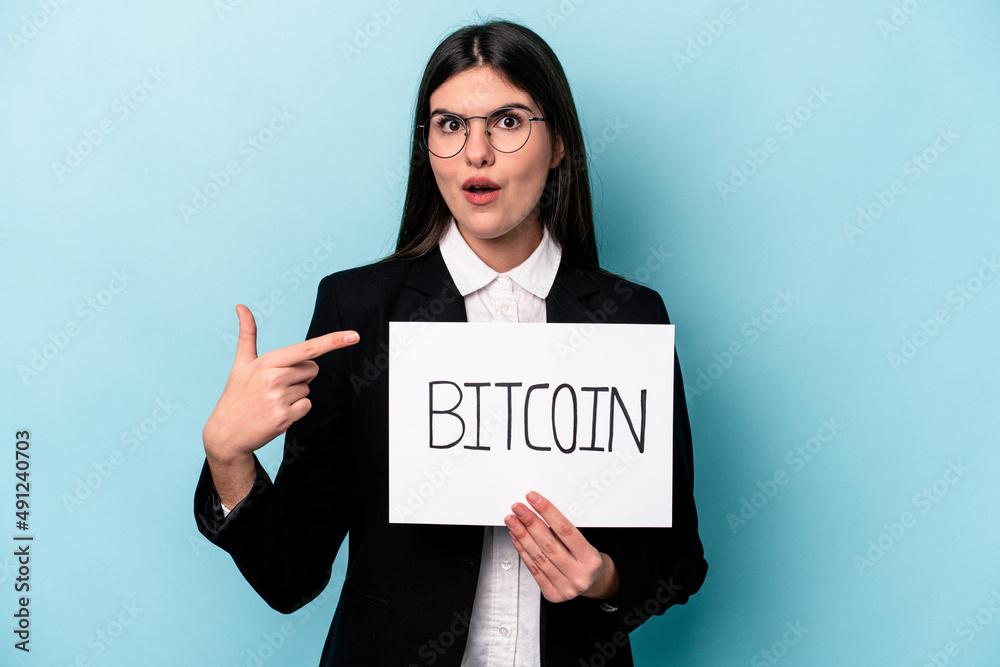 Young caucasian business woman holding a bitcoin placard isolated on blue background pointing to the side Young caucasian business woman holding a bitcoin placard isolated on blue background pointing to the side
