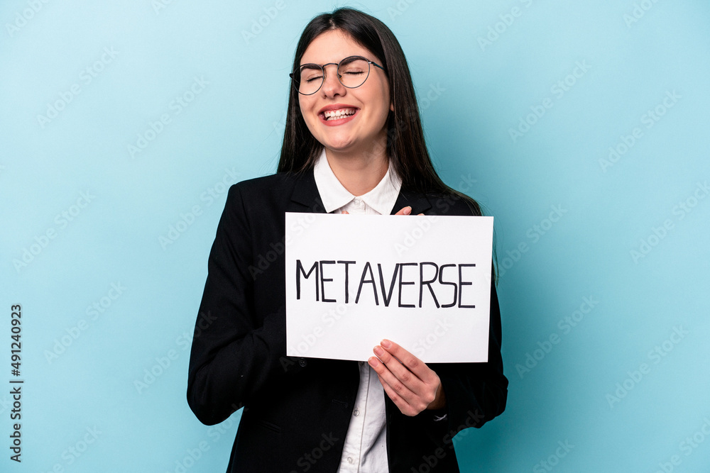 Young caucasian business woman holding a metaverse placard isolated on blue background laughs out loudly keeping hand on chest. Young caucasian business woman holding a metaverse placard isolated on blue background laughs out loudly keeping hand on chest.
