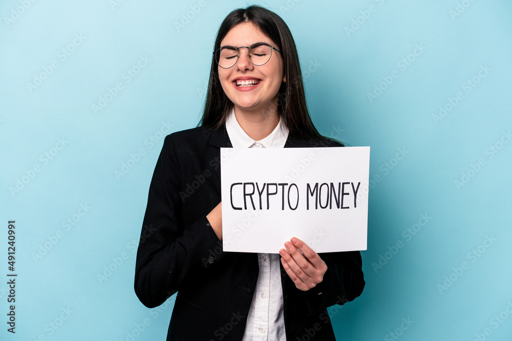 Young caucasian business woman holding a crypto money placard isolated on blue background laughs out loudly keeping hand on chest. Young caucasian business woman holding a crypto money placard isolated on blue background laughs out loudly keeping hand on chest.