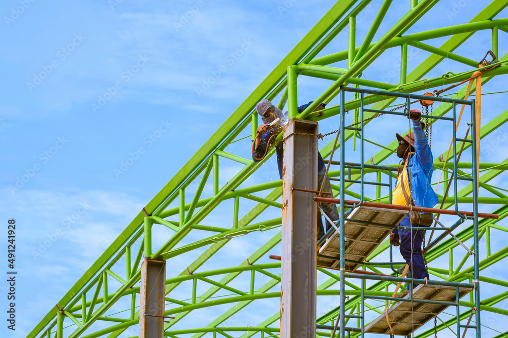 Fototapeta premium Asian foreman and construction worker on scaffolding are welding metal roof structure of industrial building against blue sky in perspective side view