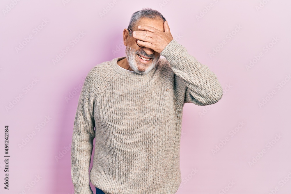 Handsome senior man with beard wearing casual sweater and glasses smiling and laughing with hand on face covering eyes for surprise. blind concept. Handsome senior man with beard wearing casual sweater and glasses smiling and laughing with hand on face covering eyes for surprise. blind concept.