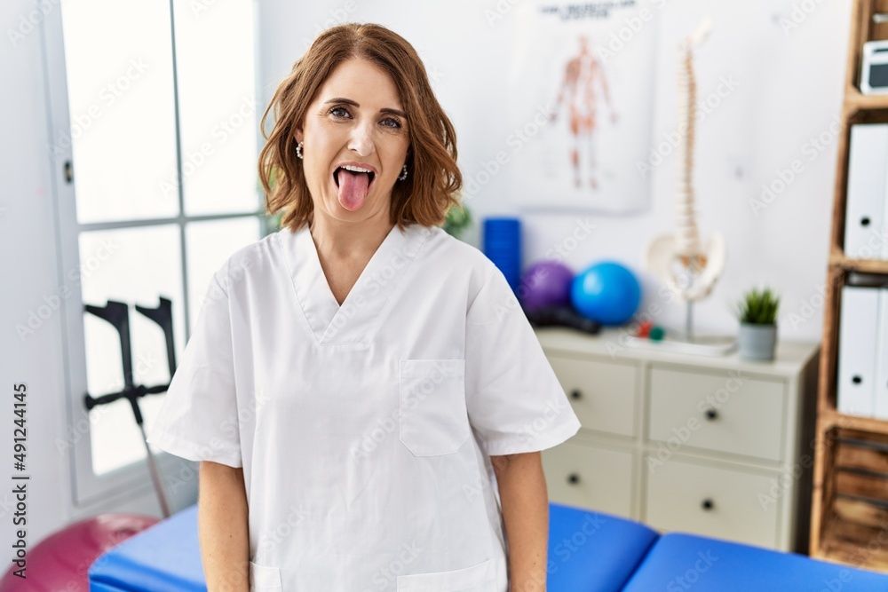 Middle age physiotherapist woman working at pain recovery clinic sticking tongue out happy with funny expression. emotion concept. Middle age physiotherapist woman working at pain recovery clinic sticking tongue out happy with funny expression. emotion concept.