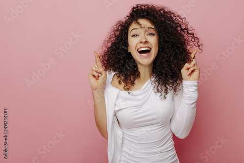 Joyful fair-skinned young girl crosses her fingers with both hands smiling broadly on pink background. Puffy-haired curly brunette in white blouse. Beauty, emotions people concept