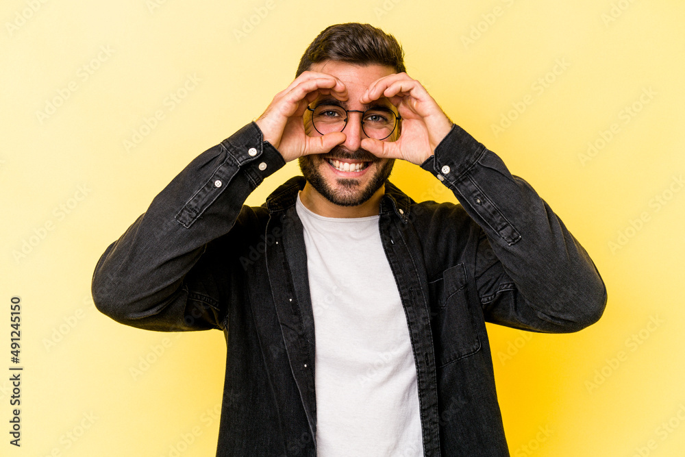 Young caucasian man isolated on yellow background showing okay sign over eyes Young caucasian man isolated on yellow background showing okay sign over eyes