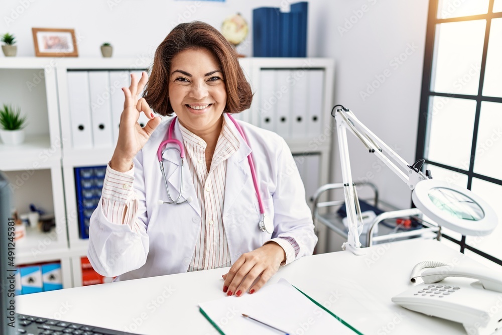 Middle age hispanic woman wearing doctor uniform and stethoscope at the clinic smiling positive doing ok sign with hand and fingers. successful expression. Middle age hispanic woman wearing doctor uniform and stethoscope at the clinic smiling positive doing ok sign with hand and fingers. successful expression.