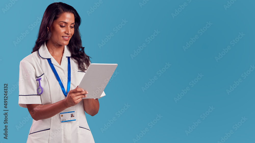 Smiling Female Mature Nurse Wearing Uniform With Digital Tablet In Front Of Blue Studio Background Smiling Female Mature Nurse Wearing Uniform With Digital Tablet In Front Of Blue Studio Background
