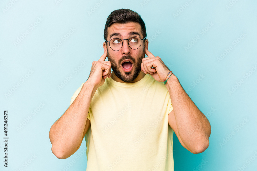 Fototapeta premium Young caucasian man isolated on blue background covering ears with fingers, stressed and desperate by a loudly ambient.