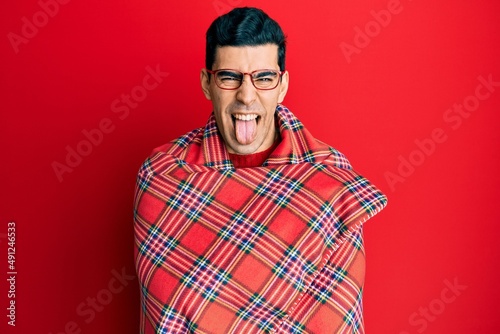 Handsome hispanic man wrapped in a red warm red blanket sticking tongue out happy with funny expression. © Krakenimages.com
