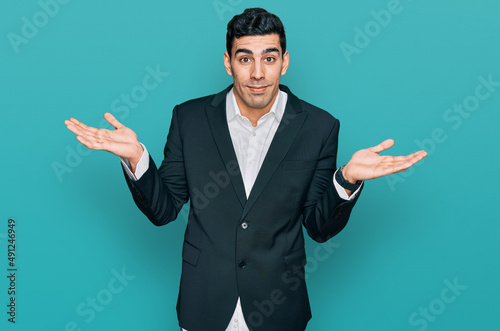 Handsome hispanic man wearing business clothes clueless and confused expression with arms and hands raised. doubt concept.