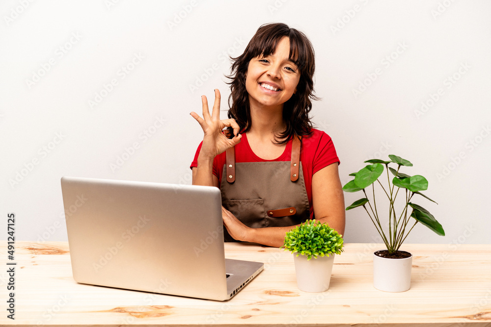Young gardener hispanic woman sitting on a table isolated on white background cheerful and confident showing ok gesture. Young gardener hispanic woman sitting on a table isolated on white background cheerful and confident showing ok gesture.