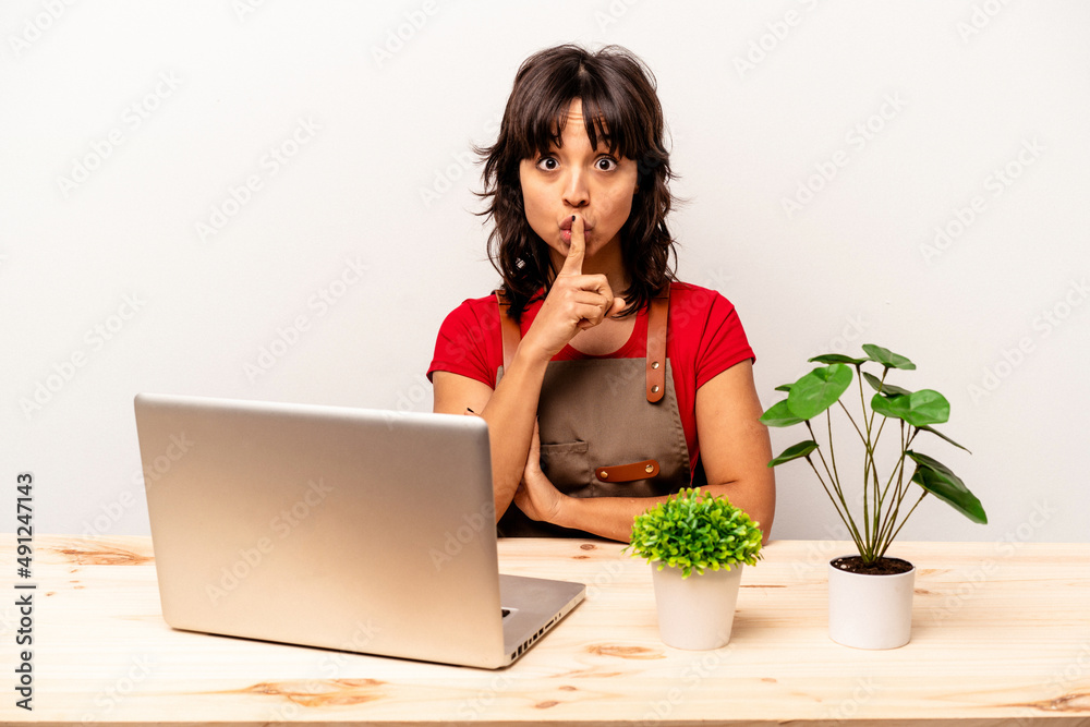 Young gardener hispanic woman sitting on a table isolated on white background keeping a secret or asking for silence. Young gardener hispanic woman sitting on a table isolated on white background keeping a secret or asking for silence.