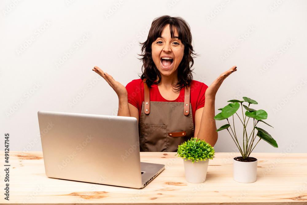 Young gardener hispanic woman sitting on a table isolated on white background receiving a pleasant surprise, excited and raising hands. Young gardener hispanic woman sitting on a table isolated on white background receiving a pleasant surprise, excited and raising hands.