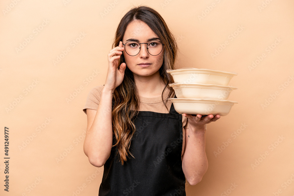 Young caucasian woman holding tupperware isolated on beige background pointing temple with finger, thinking, focused on a task. Young caucasian woman holding tupperware isolated on beige background pointing temple with finger, thinking, focused on a task.