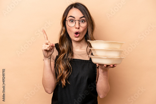 Young caucasian woman holding tupperware isolated on beige background having some great idea, concept of creativity.