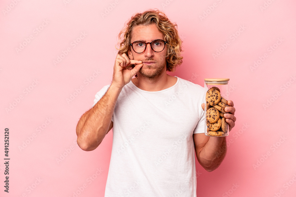 Young caucasian man holding cookies jar isolated on pink background with fingers on lips keeping a secret. Young caucasian man holding cookies jar isolated on pink background with fingers on lips keeping a secret.