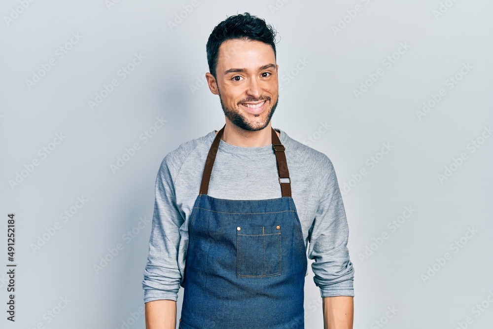 Fototapeta premium Young hispanic man wearing apron and gloves looking positive and happy standing and smiling with a confident smile showing teeth