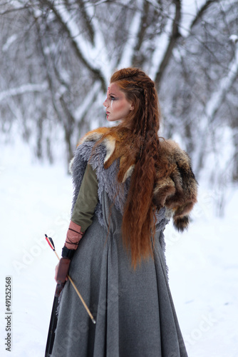 Viking warrior girl with paited face and arrow in her hand dressed in fur coat in winter forest. Fantasy portrait of woman
