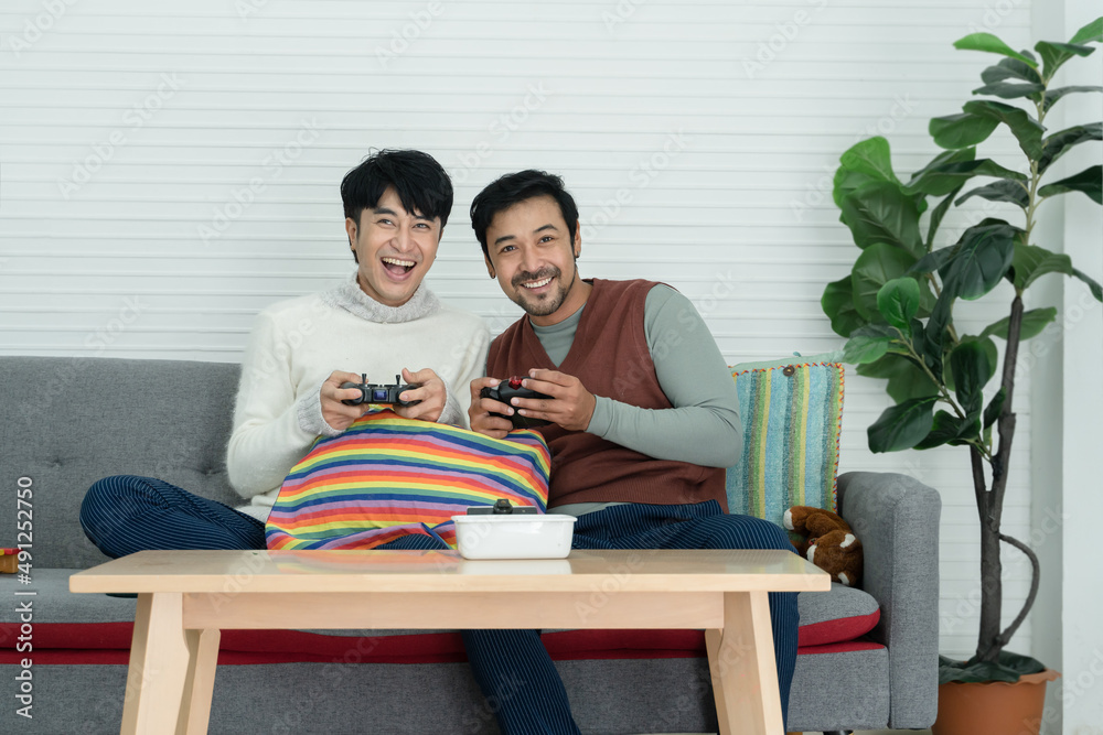 Happy Asian young gay couple wearing sweater holding joysticks playing game with excited face have fun together at living room at home. Lifestyle of Lgbt family in holiday