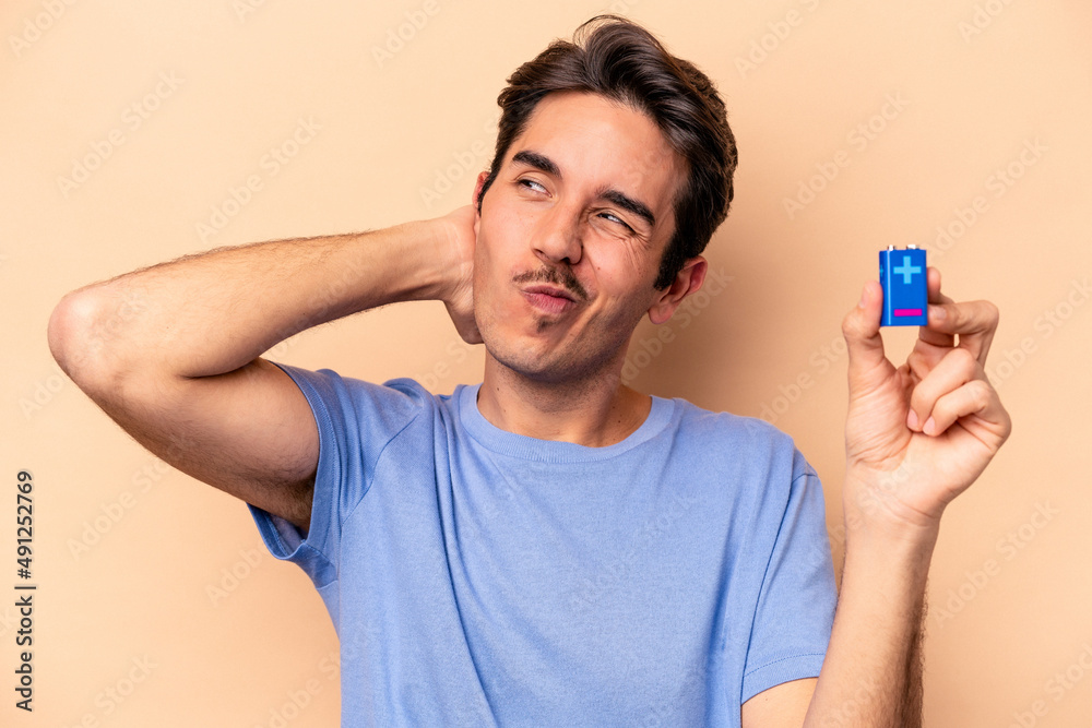 Young caucasian man holding a batterie isolated on beige background touching back of head, thinking and making a choice.