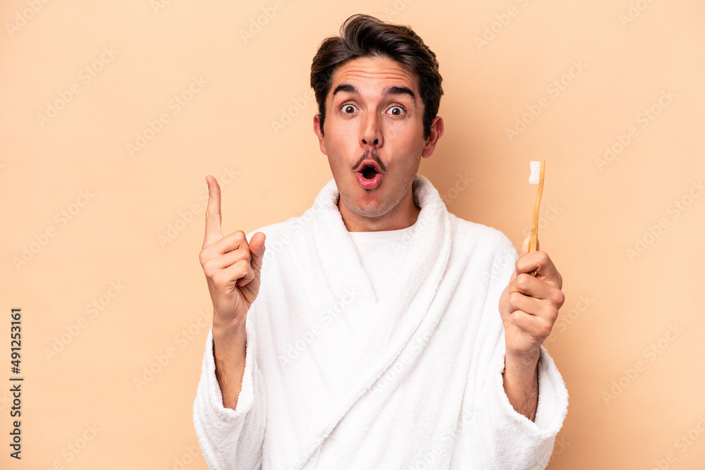 Young caucasian man wearing a bathrobe holding a toothbrush isolated on beige background having some great idea, concept of creativity.