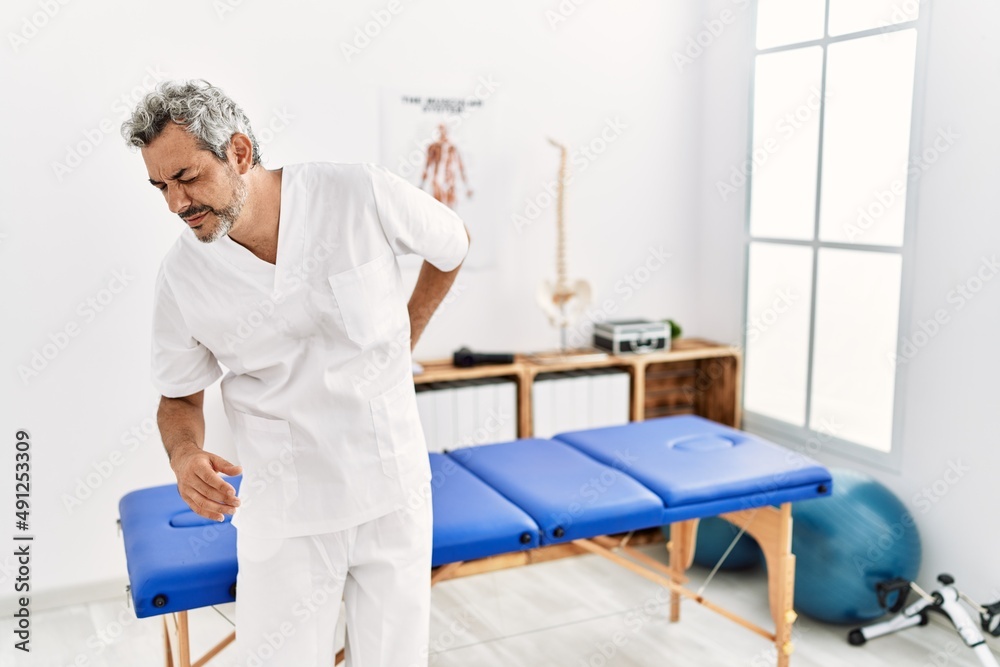Middle age hispanic therapist man working at pain recovery clinic suffering of backache, touching back with hand, muscular pain