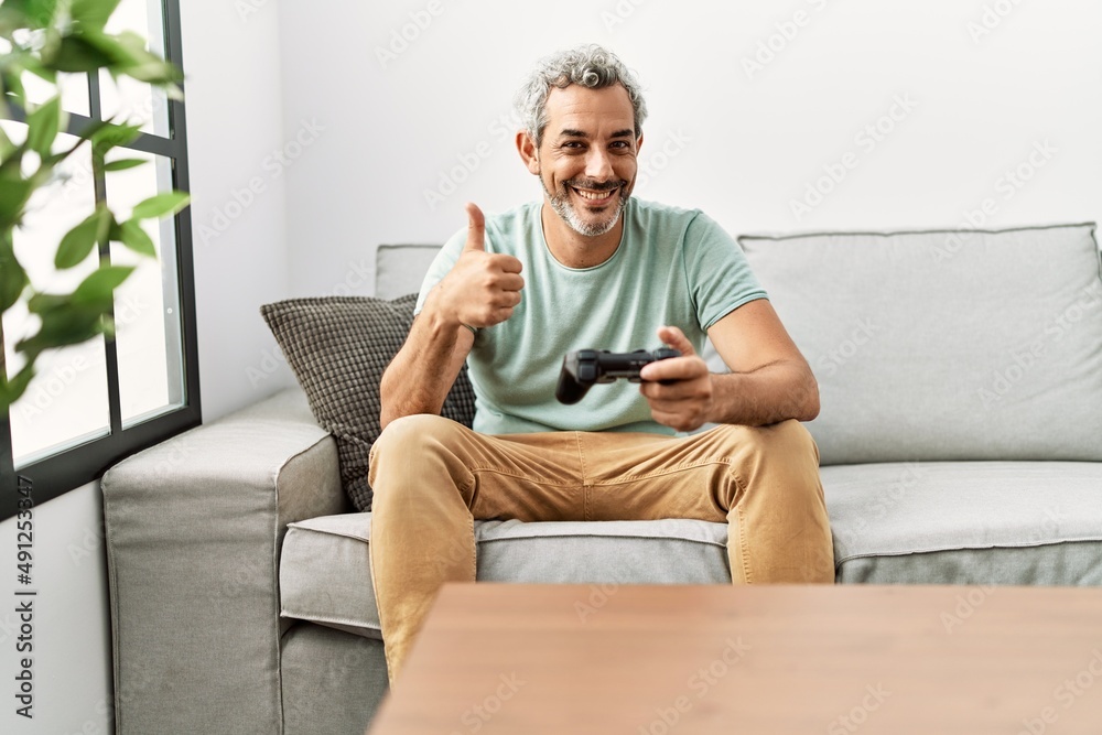 Middle age hispanic man playing video game sitting on the sofa smiling happy and positive, thumb up doing excellent and approval sign