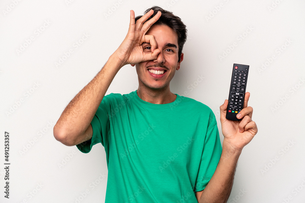 Young caucasian man holding a tv remote isolated on white background excited keeping ok gesture on eye. Young caucasian man holding a tv remote isolated on white background excited keeping ok gesture on eye.