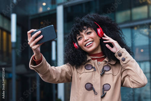 Cheerful woman taking listening music through wireless headphones taking selfie through smart phone in city photo