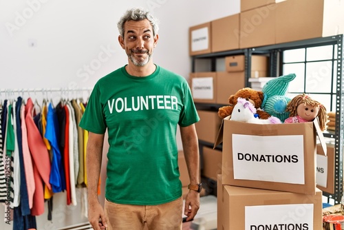 Middle age hispanic man wearing volunteer t shirt at donations stand smiling looking to the side and staring away thinking.
