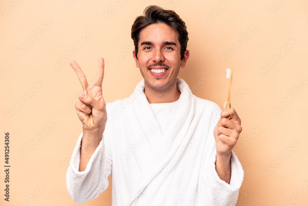 Young caucasian man wearing a bathrobe holding a toothbrush isolated on beige background showing number two with fingers. Young caucasian man wearing a bathrobe holding a toothbrush isolated on beige background showing number two with fingers.