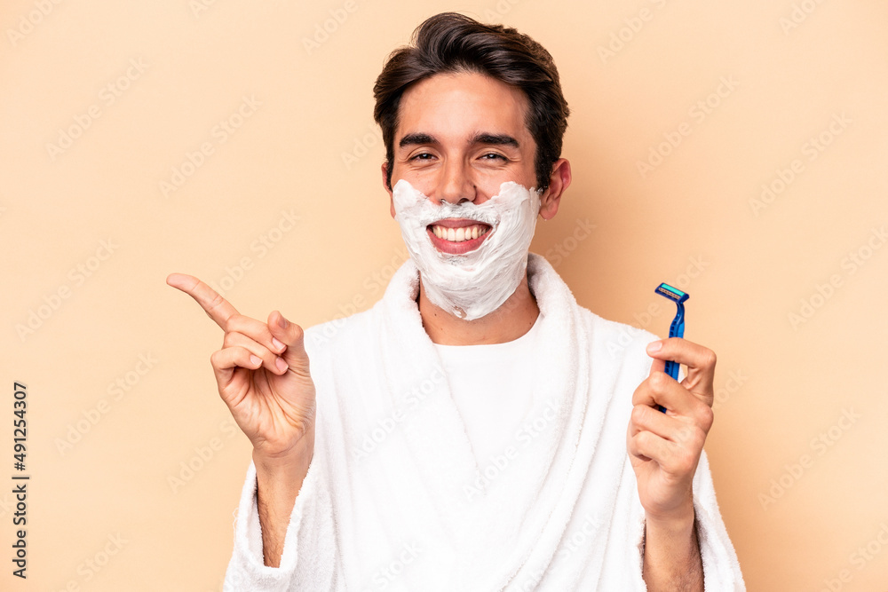 Young caucasian man shaving his beard isolated on beige background smiling and pointing aside, showing something at blank space.