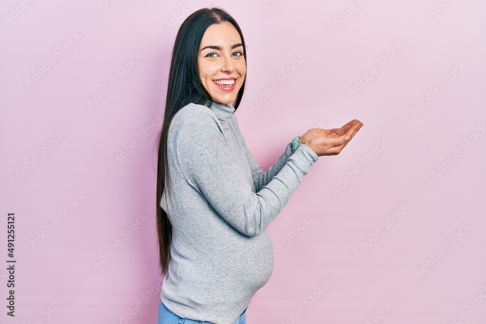 Beautiful woman with blue eyes expecting a baby, touching pregnant belly pointing aside with hands open palms showing copy space, presenting advertisement smiling excited happy