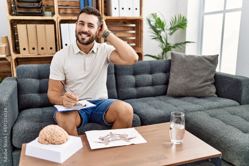 Handsome hispanic man working with rorschach test at psychology clinic smiling confident touching hair with hand up gesture, posing attractive and fashionable Handsome hispanic man working with rorschach test at psychology clinic smiling confident touching hair with hand up gesture, posing attractive and fashionable