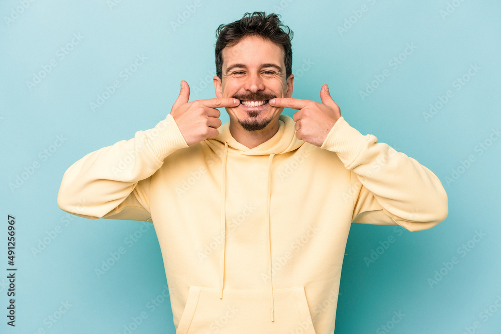 Fototapeta premium Young caucasian man isolated on blue background smiles, pointing fingers at mouth.