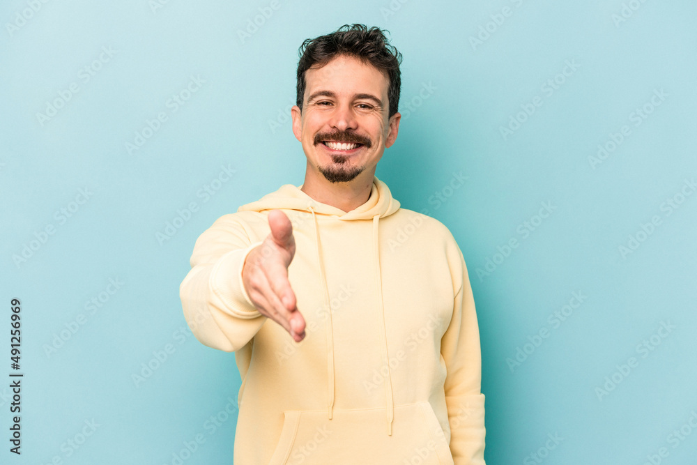Young caucasian man isolated on blue background stretching hand at camera in greeting gesture. Young caucasian man isolated on blue background stretching hand at camera in greeting gesture.