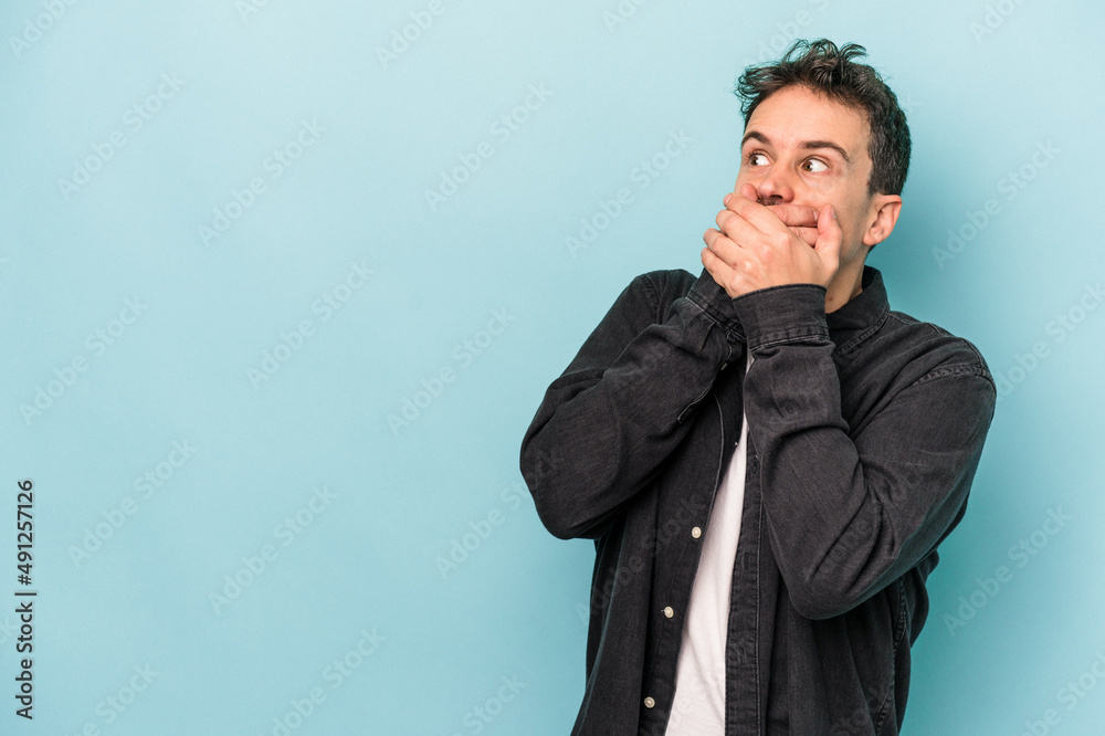Young caucasian man isolated on blue background thoughtful looking to a copy space covering mouth with hand.
