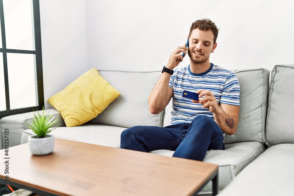 Young hispanic man talking on the smartphone holding credit card at home