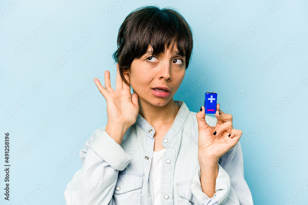 Young hispanic woman holding a batterie isolated on blue background trying to listening a gossip. Young hispanic woman holding a batterie isolated on blue background trying to listening a gossip.