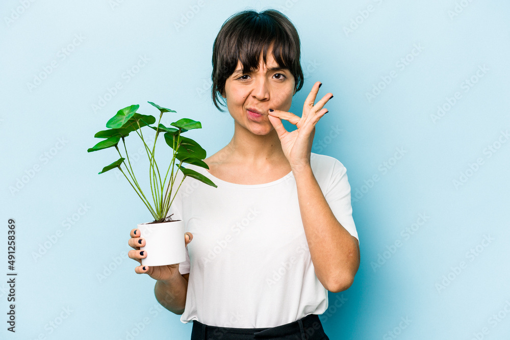 Young hispanic woman holding a plant isolated on blue background with fingers on lips keeping a secret. Young hispanic woman holding a plant isolated on blue background with fingers on lips keeping a secret.